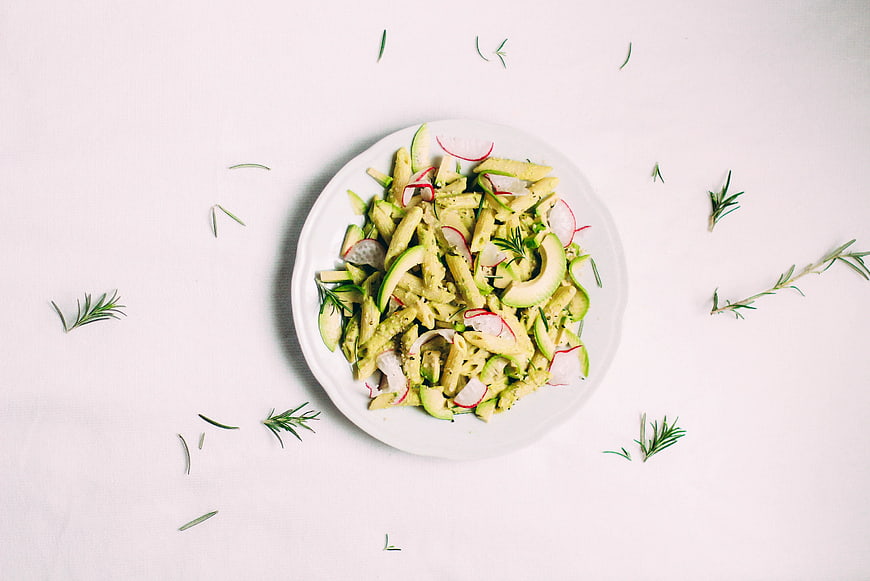 Top view of penne pasta with pesto, avocado, and radishes on white background
