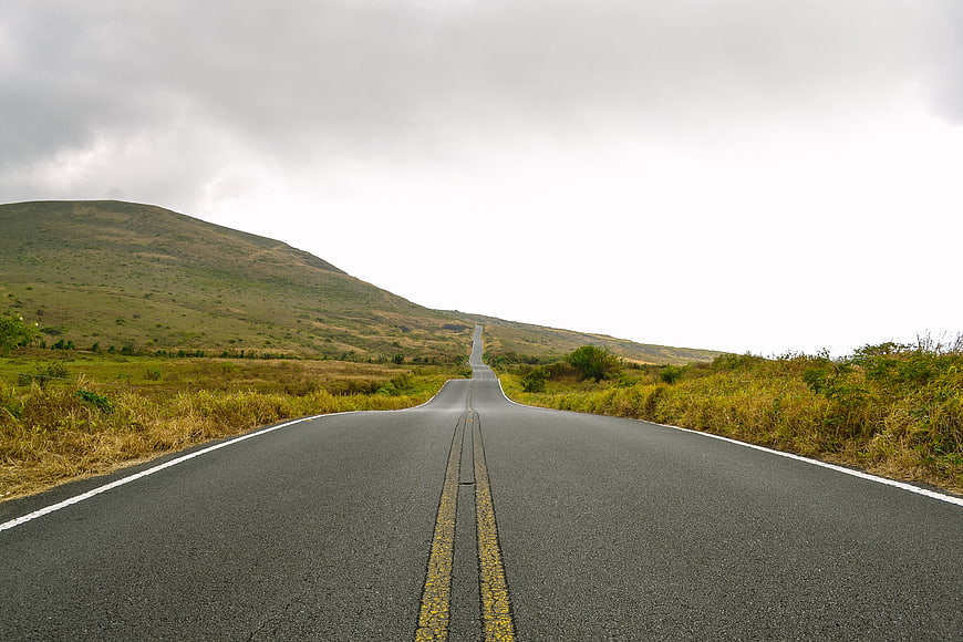 Empty asphalt road winding through grassy hills under cloudy sky