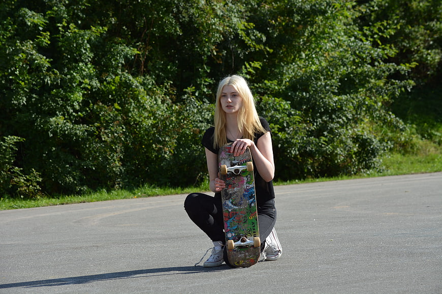 Blonde Woman Crouching with Skateboard on Asphalt Road