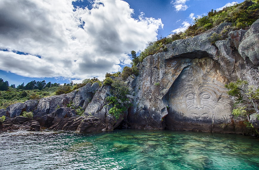 Maori Rock Carving Face In Coastal Cliff With Turquoise Water