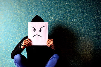Person holding angry face paper mask against blue wall