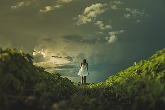 Woman in White Dress Walking Through Green Field Under Dramatic Clouds