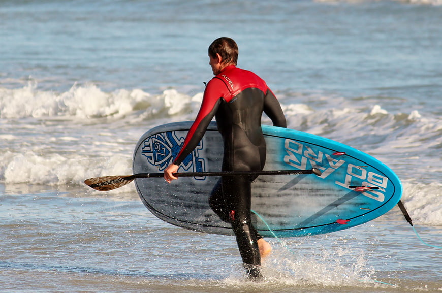 Man in Wetsuit Carrying Stand Up Paddleboard into Ocean
