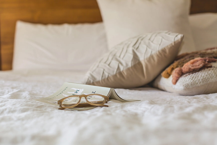 Reading glasses and book on cozy white bed with pillows