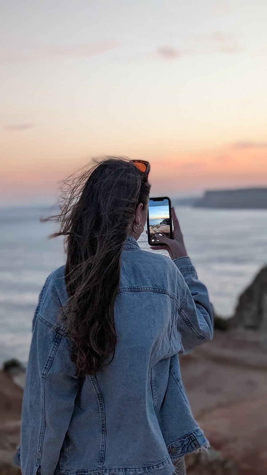 Woman photographing sunset ocean view with smartphone from rocky coast
