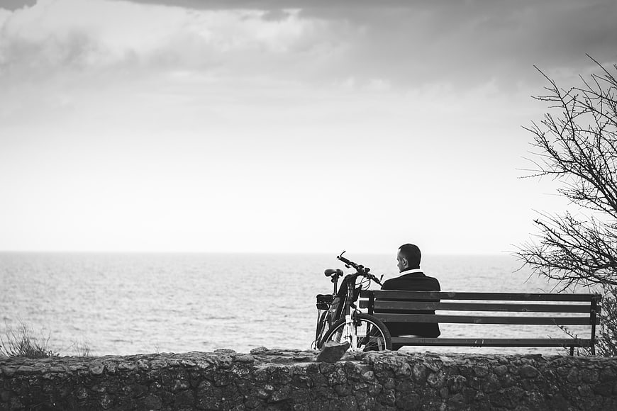 Man sitting on bench by the sea with bicycle in black and white