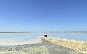 Red SUV and lone figure on salt flat road under blue sky