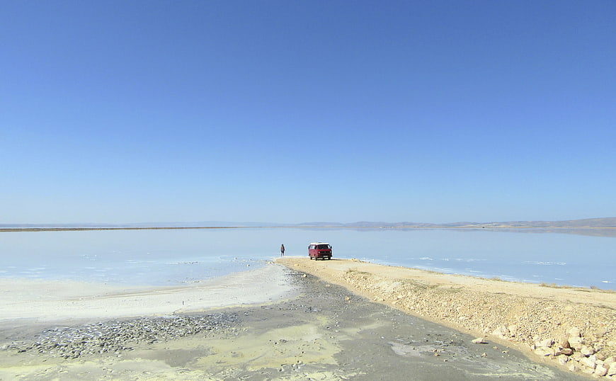 Red SUV and lone figure on salt flat road under blue sky
