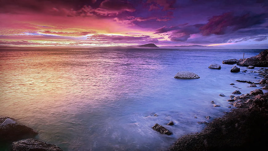 Vibrant purple and pink sunset over calm ocean with rocky shore