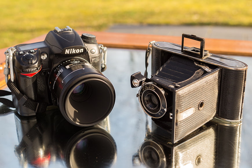 Modern Nikon DSLR and vintage folding camera on glass table