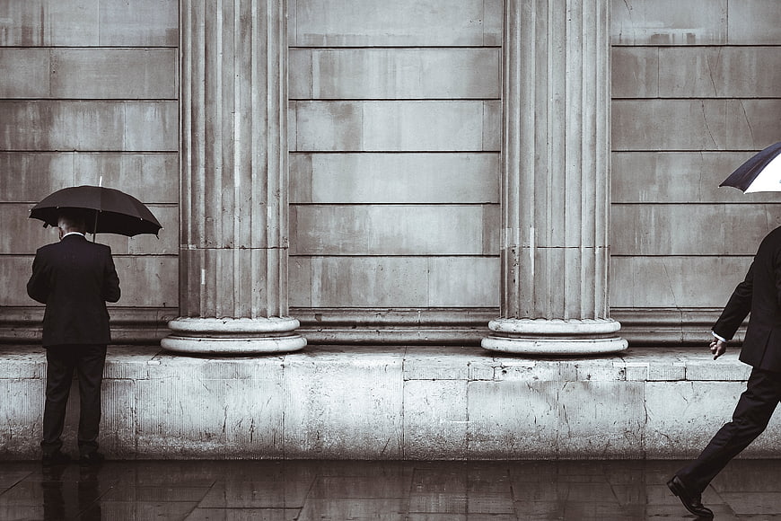 Two Men with Umbrellas Walking Past Classical Columns in Rain