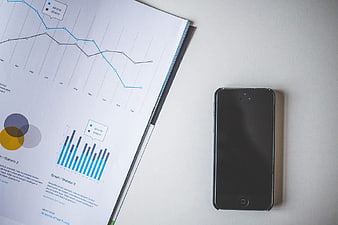 Black smartphone next to financial report charts on white desk