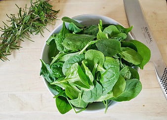 Fresh spinach leaves in white bowl with rosemary sprigs and knife on wooden surface