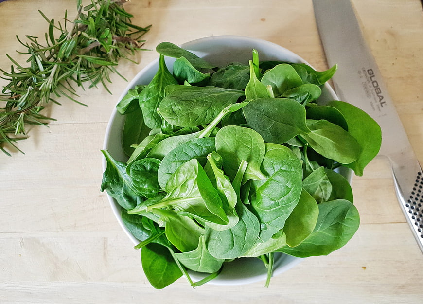 Fresh spinach leaves in white bowl with rosemary sprigs and knife on wooden surface