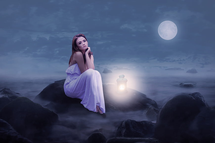 Woman in white dress sitting on rocks with lantern under full moon