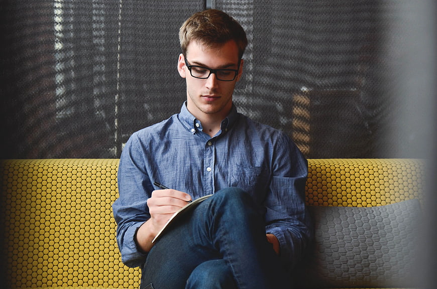 Young man with glasses writing in notebook on yellow couch
