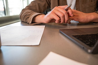 Close-up of hands holding pen at desk with laptop and documents