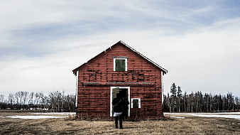 Person standing before weathered red wooden house in snowy field