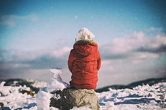 Child in Red Jacket Sitting on Snowy Rock Looking at Sky