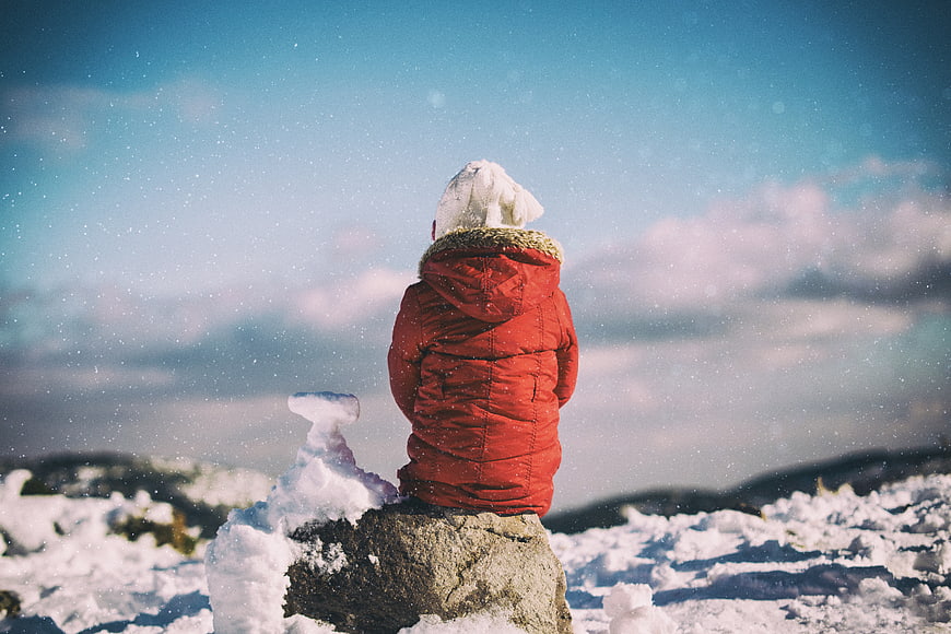 Child in Red Jacket Sitting on Snowy Rock Looking at Sky