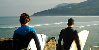 Two surfers in wetsuits looking at ocean waves