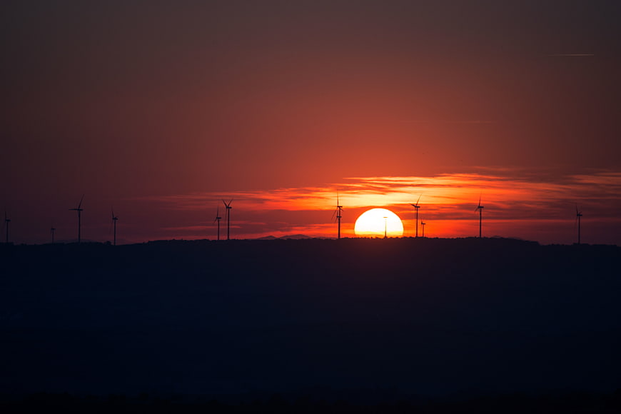 Silhouette of wind turbines on a hill against a vibrant orange sunset sky