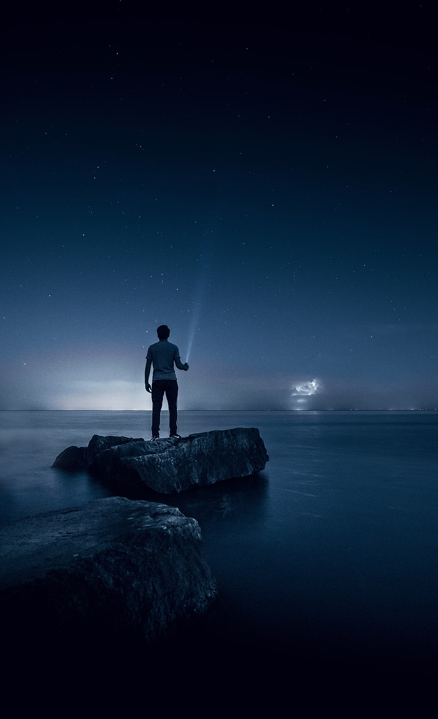 Man standing on rock at night with flashlight under starry sky