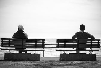 Two people sitting on separate benches by the water in black and white
