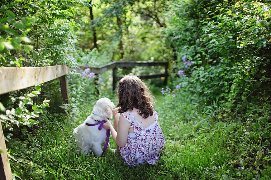 Little girl sitting with white puppy in lush green garden