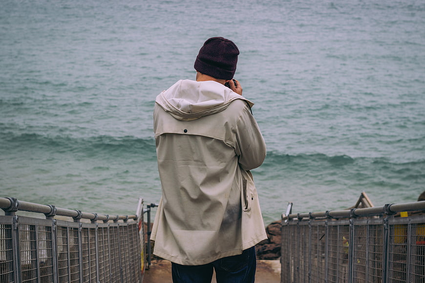 Man in Beige Coat Looking at Ocean from Pier