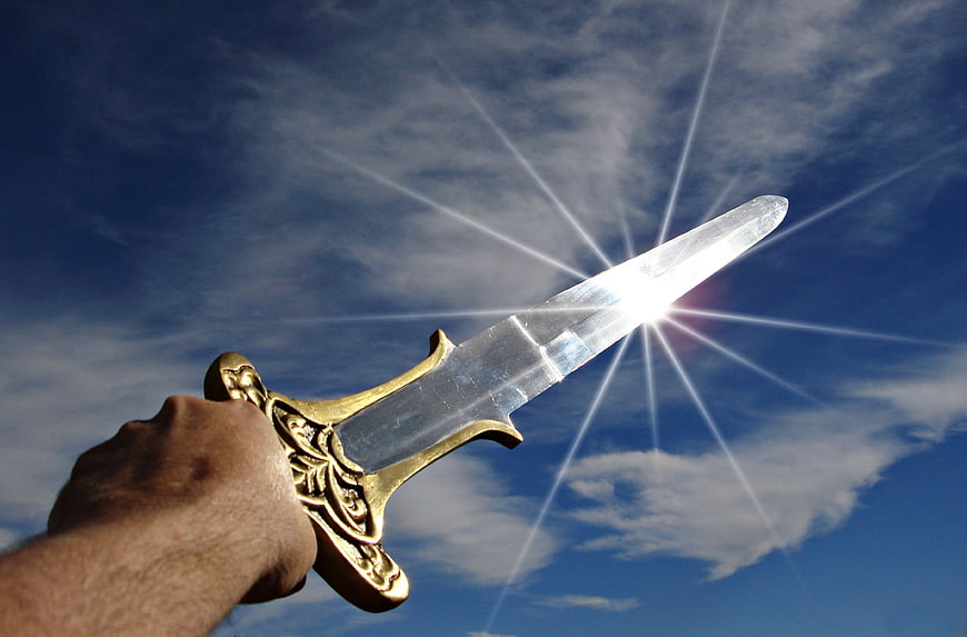 Hand holding golden sword against blue sky with sun flare