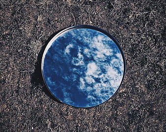 Round mirror reflecting blue sky and clouds on dry grass