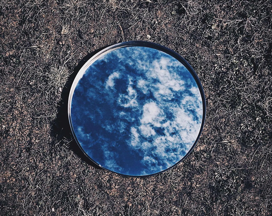 Round mirror reflecting blue sky and clouds on dry grass