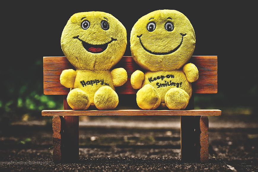Two yellow smiley face plush toys sitting on a wooden bench