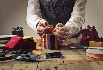 Man Wrapping Gift Box with Orange Ribbon on Wooden Table