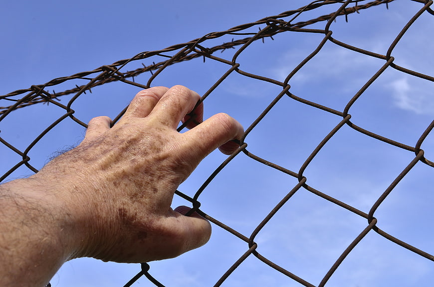 Man's Hand Gripping Chain Link Fence With Barbed Wire Against Blue Sky