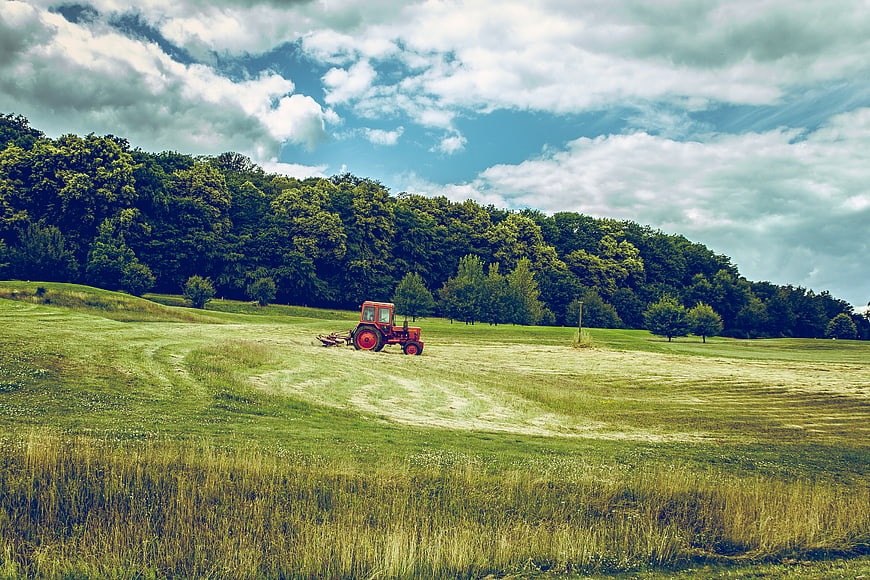 Red Tractor Mowing Green Pasture Under Cloudy Sky