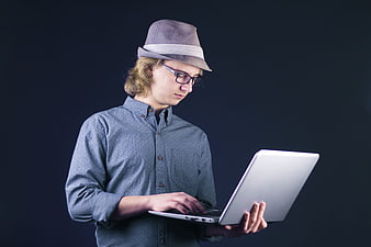 Young man in fedora and glasses using a laptop against a dark background