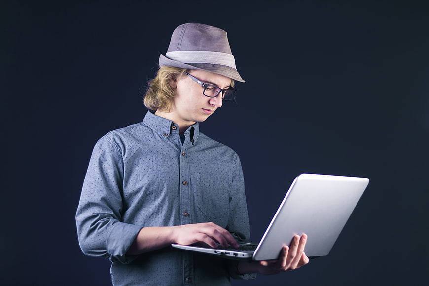 Young man in fedora and glasses using a laptop against a dark background
