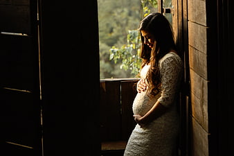 Pregnant woman in white lace dress holding belly by window
