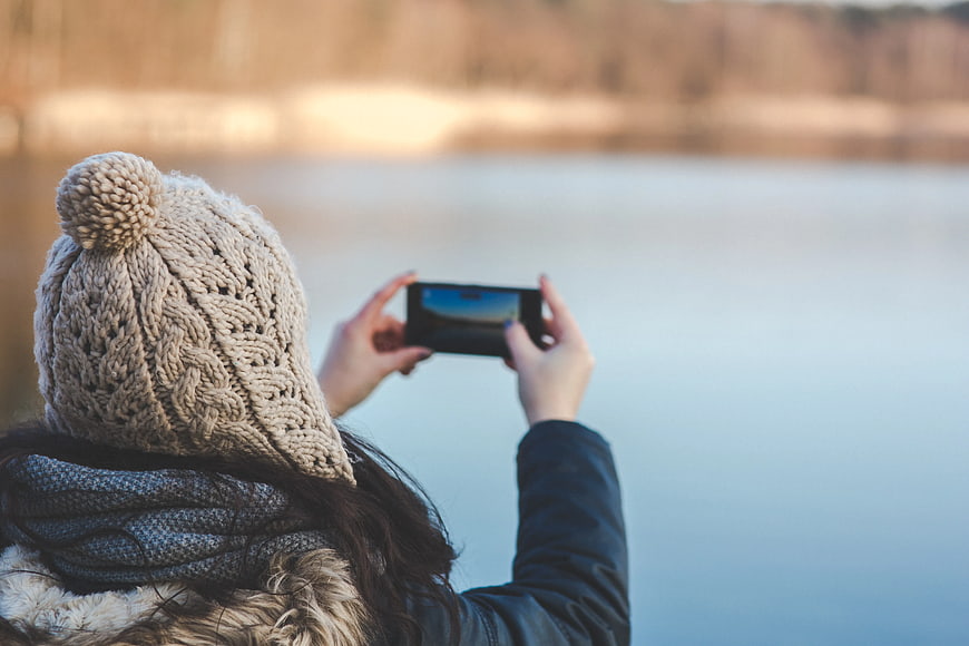 Woman in winter hat taking photo of lake with smartphone