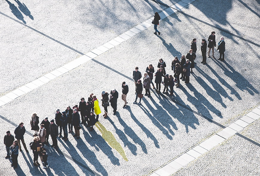 Aerial view of people queuing on cobblestone plaza with long shadows