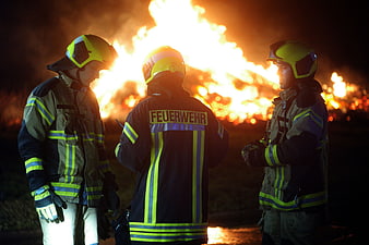Three German Firefighters Standing Before Large Night Fire