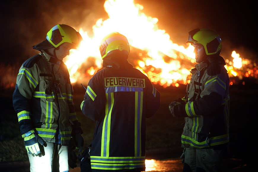 Three German Firefighters Standing Before Large Night Fire