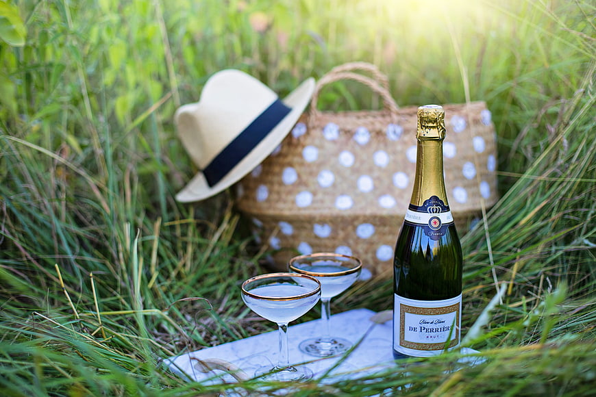 Champagne Picnic Setup With Straw Hat And Wicker Basket In Summer Grass