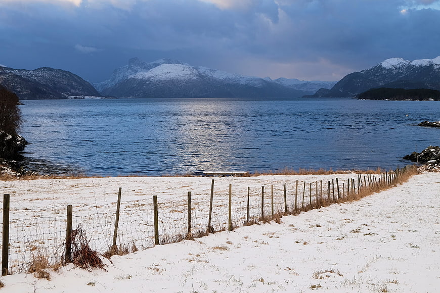 Snowy coastal landscape with wooden fence and fjord mountains
