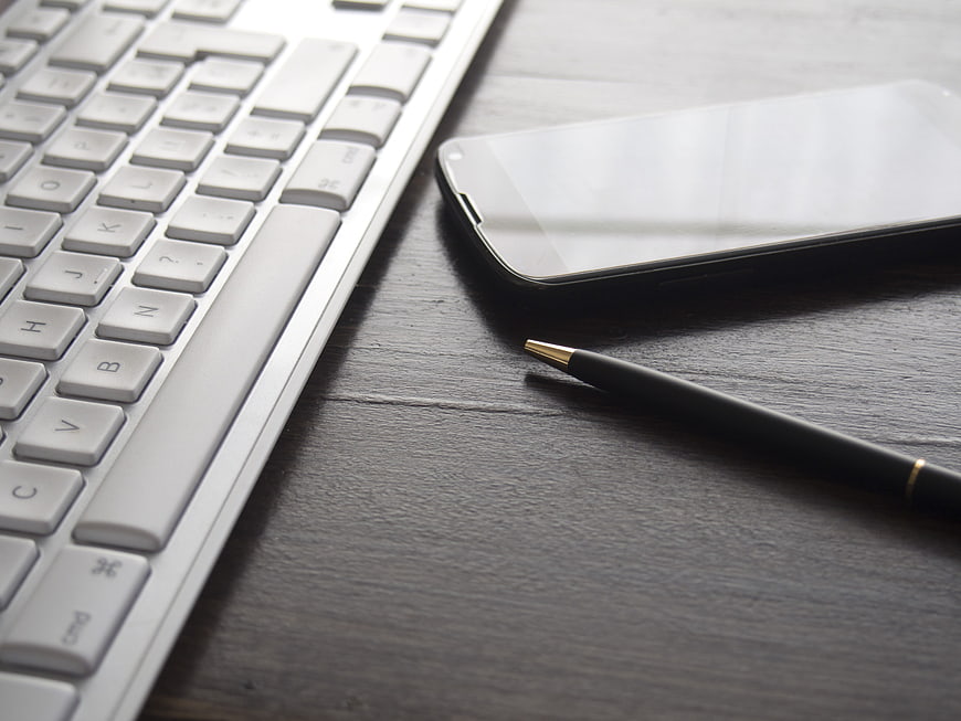 White keyboard, smartphone, and pen on dark wooden desk
