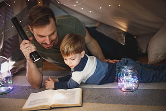 Father and son reading book in blanket fort with fairy lights