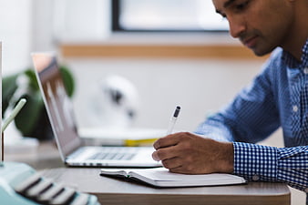 Man writing in notebook at office desk with laptop