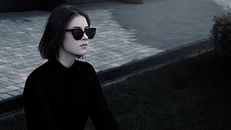Young woman in black sunglasses and dark top sitting on a city street